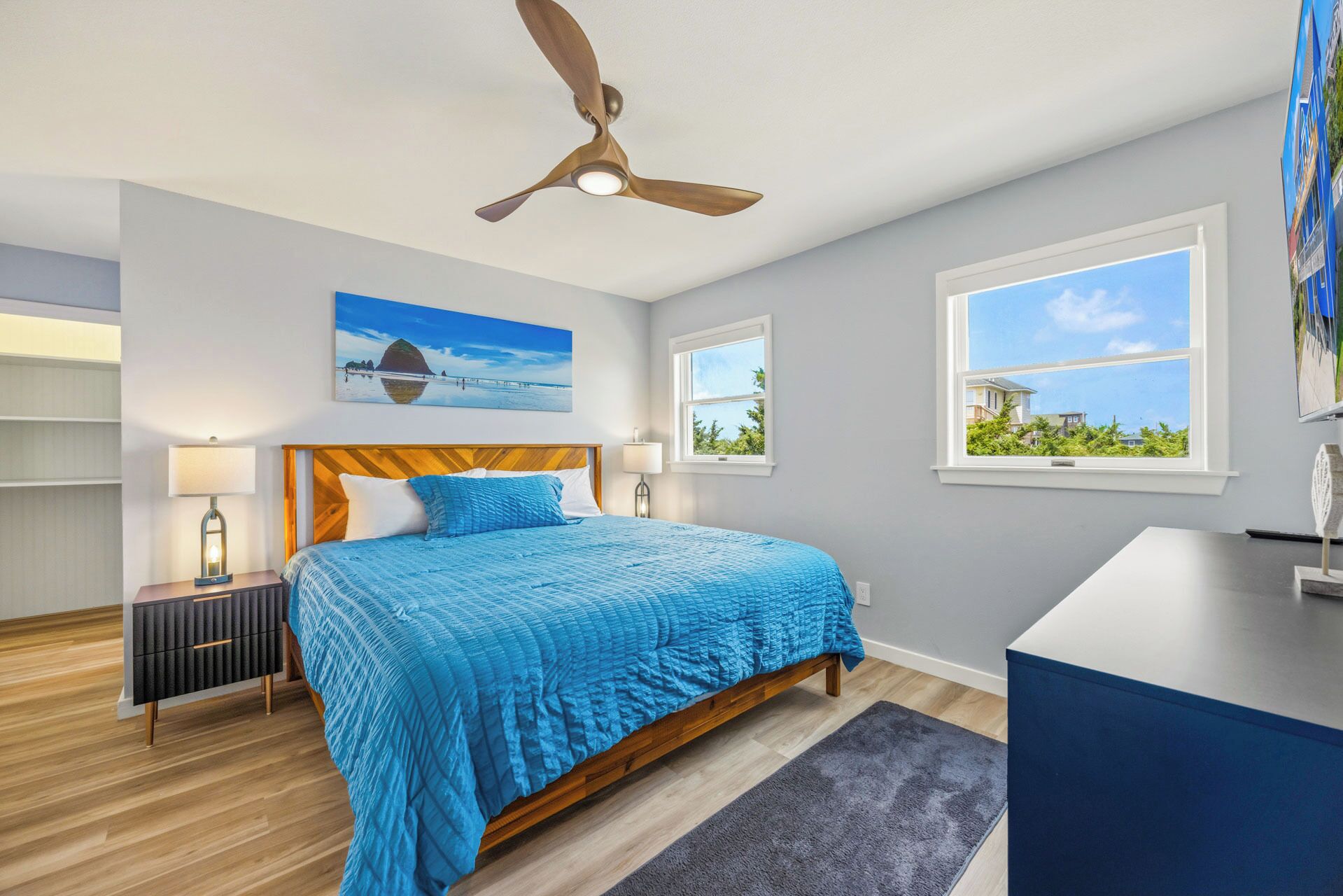 A bedroom with a bed, a ceiling fan, a nightstand, a lamp, a painting, and a window with a view.