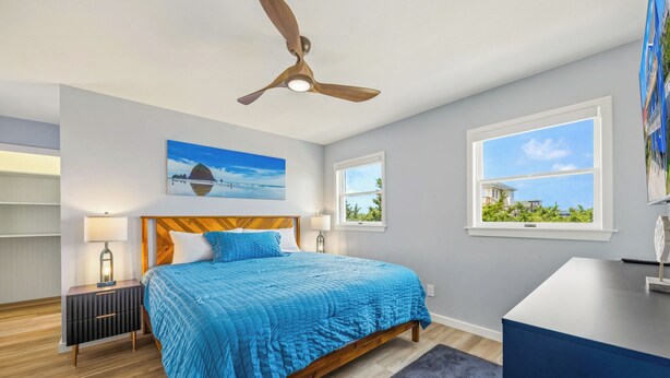 A bedroom with a bed, a ceiling fan, a nightstand, a lamp, a painting, and a window with a view. - Brand New Semi-Oceanfront Waves Home with Private Pool, Hot Tub and Game Room (Waves)