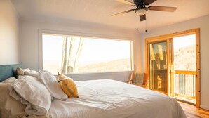 Un dormitorio con una cama grande, ventilador de techo, ventana con vistas a árboles y puerta corrediza de vidrio a una terraza.