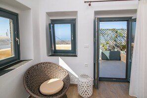Habitación con una silla de mimbre, una mesita pequeña, un cubo de basura blanco y un ventanal corredizo que da a un espacio exterior con vista al mar.