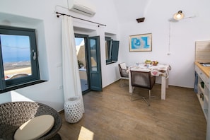 Una habitación con zona de comedor, una silla de mimbre, una mesa con mantel blanco y vista al mar a través de una ventana.