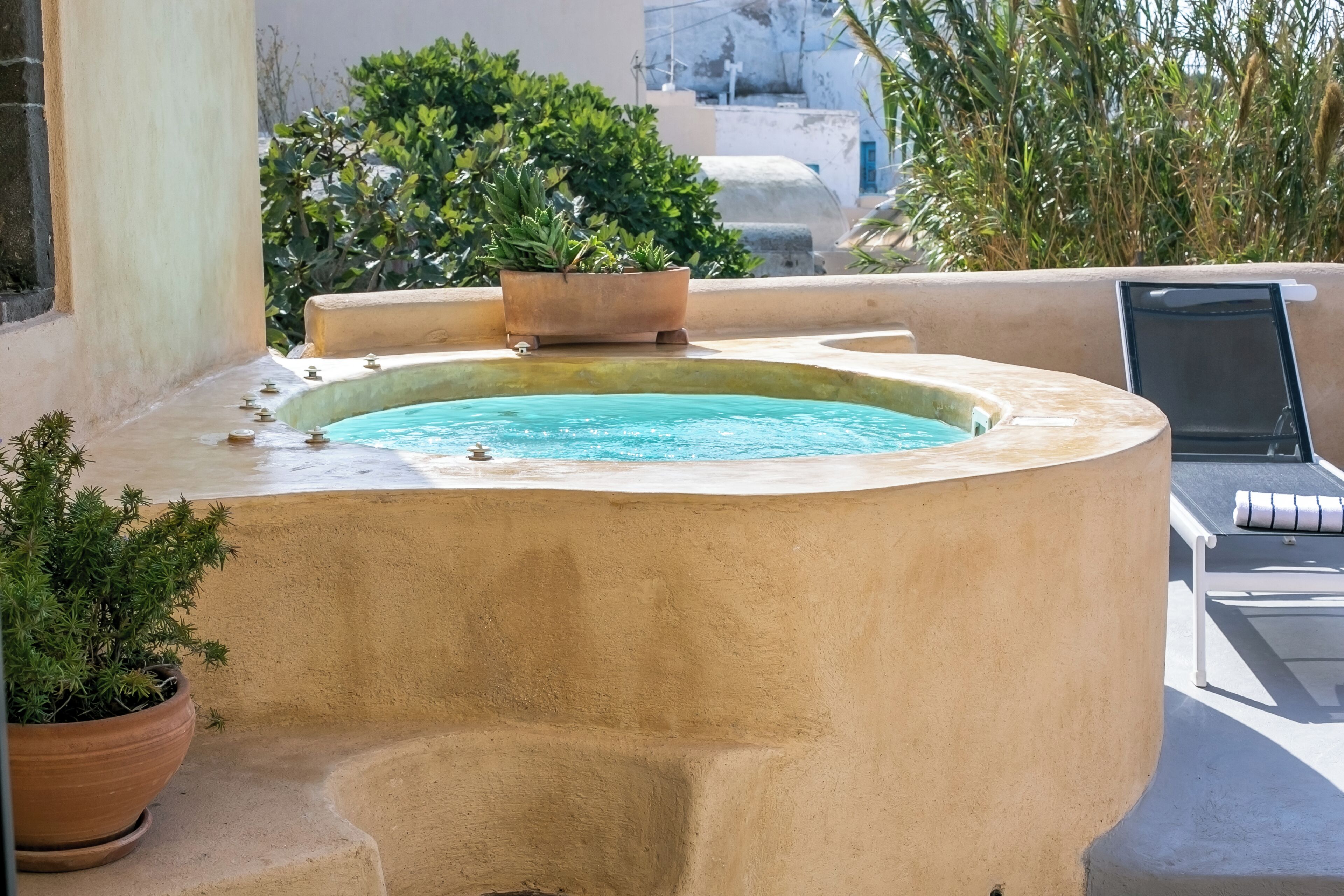 A hot tub with a curved edge, surrounded by potted plants and a lounge chair.