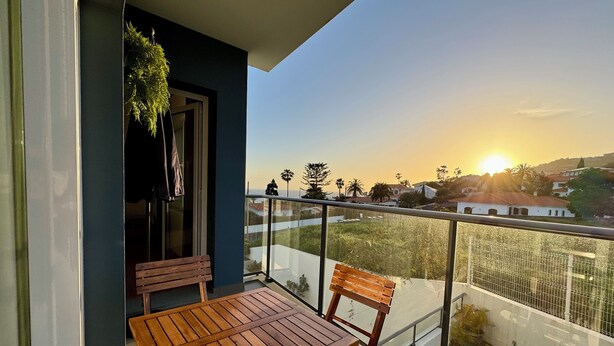 Ein Balkon mit Holztisch und -stühlen, einer hängenden Pflanze und Blick auf eine Wohngegend bei Sonnenuntergang.