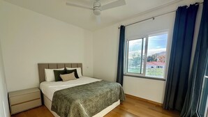 Ein Schlafzimmer mit einem Bett, einem Nachttisch, einem Deckenventilator, einem Fenster mit Vorhängen und Blick auf ein Gebäude.