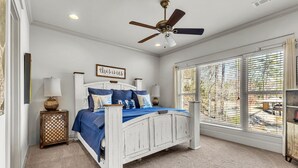 Un dormitorio con cama blanca, ropa de cama azul, ventilador de techo y una ventana grande con persianas.