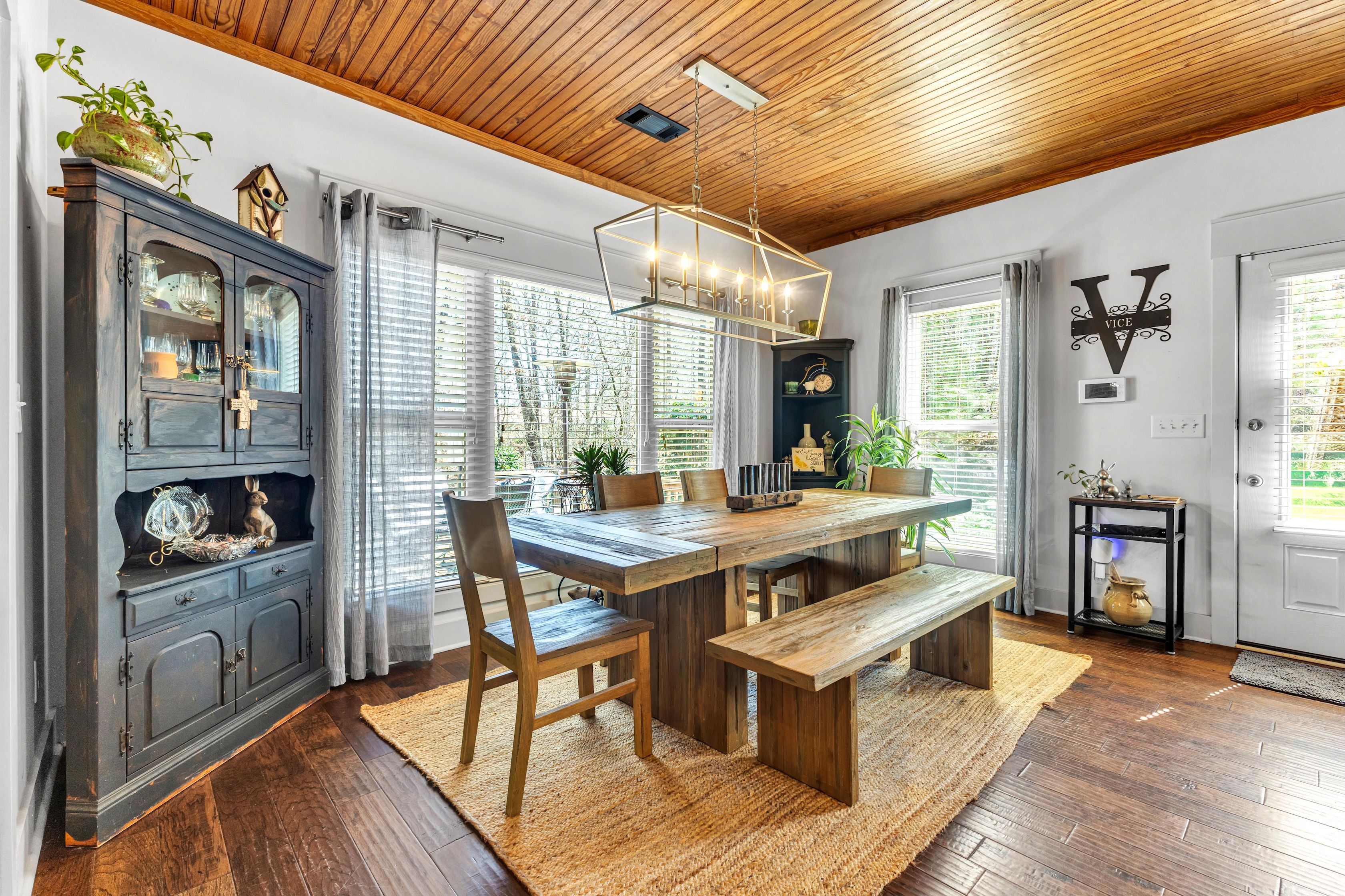 A dining area with a wooden table, chairs, and a bench. There is a dark wooden cabinet with glass doors and a small shelf. A decorative birdhouse is on top of the cabinet. The room has a wooden ceiling and a large window with blinds.