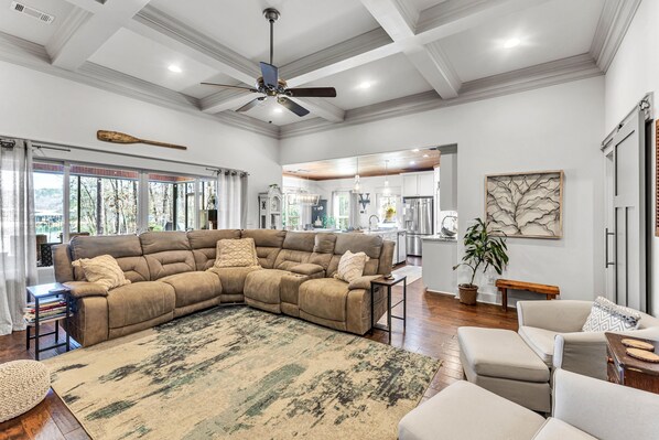 A spacious living room with a large sectional sofa, a ceiling fan, and a view of the kitchen through large windows.