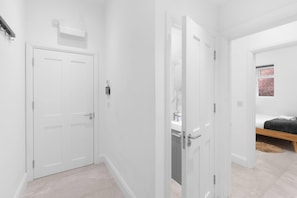 A hallway with a closed door, a white wall, and a window with a brick wall visible through it. - Modern 4 Bedroom Apartment in London with Parking + Garden (London)