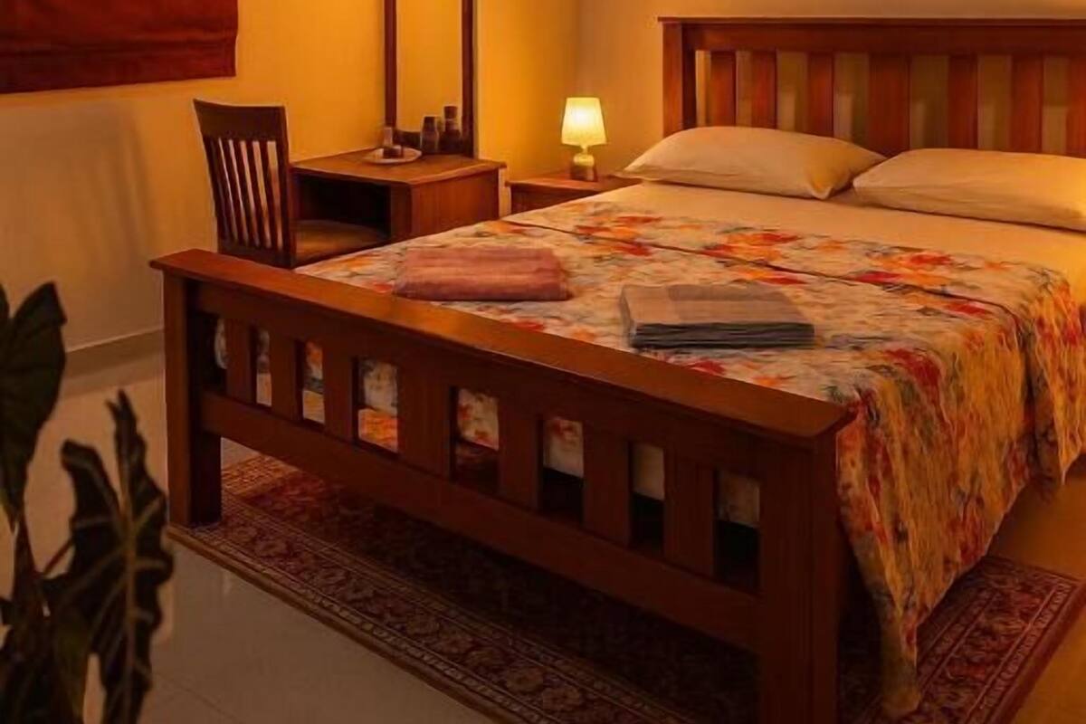 A bedroom with a wooden bed, a ceiling fan, a bedside table with a lamp, a mirror, and a potted plant.