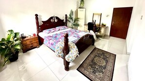 A bedroom with a wooden ceiling, a bed with a floral bedspread, a nightstand, a dresser, a mirror, a door, and a potted plant.