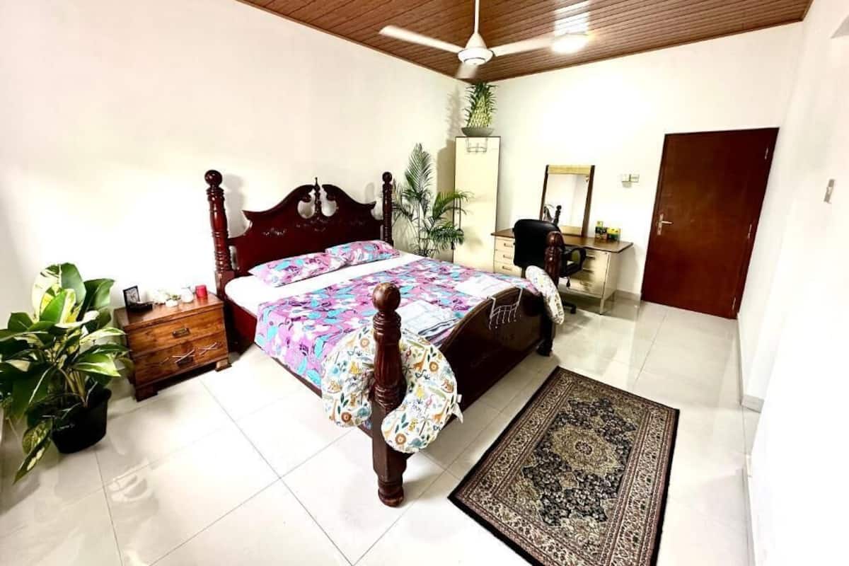 A bedroom with a wooden ceiling, a bed with a floral bedspread, a nightstand, a dresser, a mirror, a door, and a potted plant.