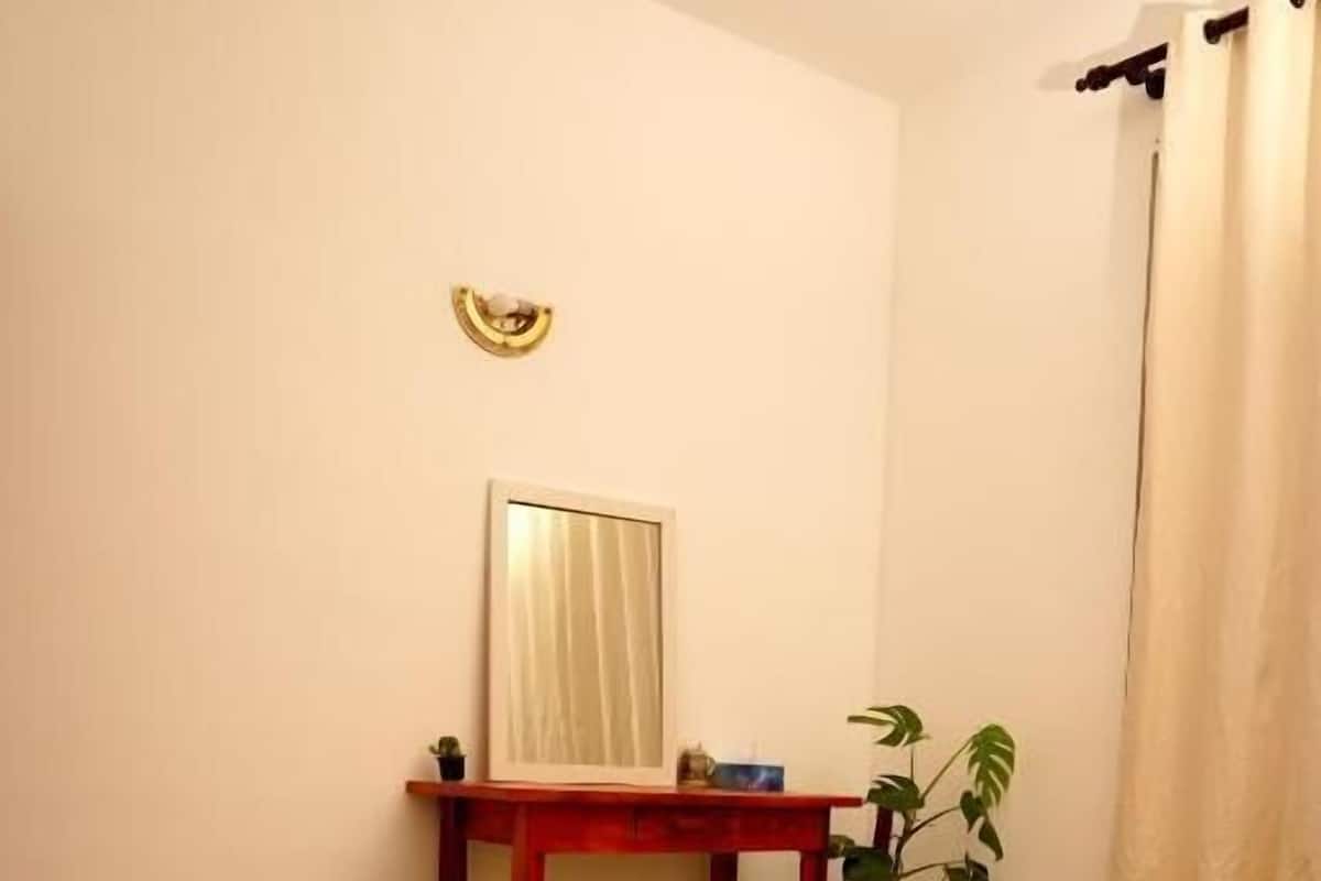 A bedroom with a bed, a bedside table, a mirror, a small plant, and a window with curtains.