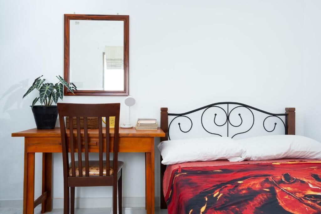 Ein Schlafzimmer mit einem Holztisch, einem Stuhl, einem Bett mit rot-schwarzem Muster sowie einem weißen Kissen und einem Spiegel an der Wand.