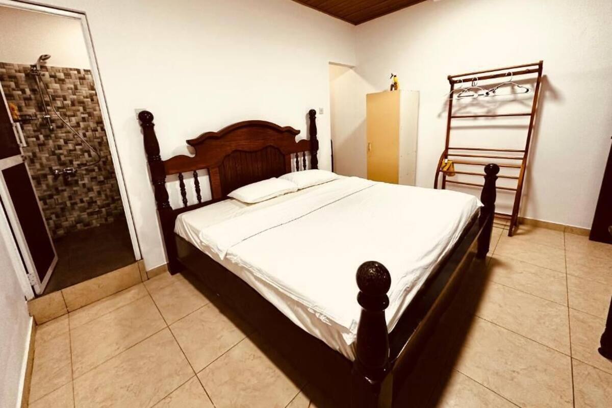 A bedroom with a wooden bed, a ceiling fan, a shower area, and a clothes drying rack.