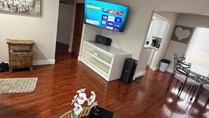 Ein Wohnzimmer mit einem an der Wand befestigten Flachbildfernseher, einem Korbwicker-Korb mit Blumen, einer weißen Unterhaltungsanlage und einem Essbereich mit Stuhl und Tisch.