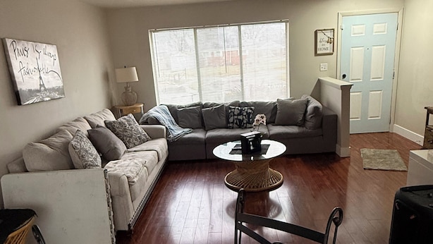 Ein Wohnzimmer mit grauem Sofa, einem runden Couchtisch und einem weißen Sessel. Es gibt ein Fenster mit Jalousien, eine Tür und einen an der Wand befestigten Bilderrahmen.