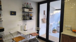 A kitchen with a white table, a chair, a shelf with jars, a picture frame, and a sliding glass door leading to a balcony with plants. - House with pool 1 km from Sitges beach (Sant Pere de Ribes)