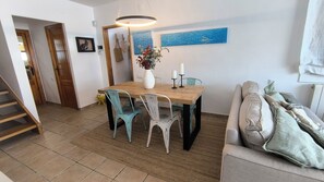 A dining area with a wooden table, four chairs, a vase with flowers, and a painting of the sea on the wall.