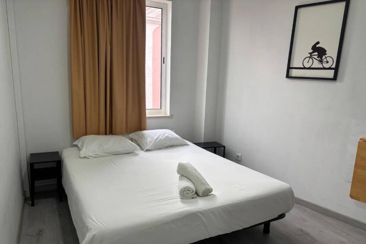 A single bed with white bedding, a rolled towel, a window with brown curtains, a framed picture on the wall, and a small nightstand.