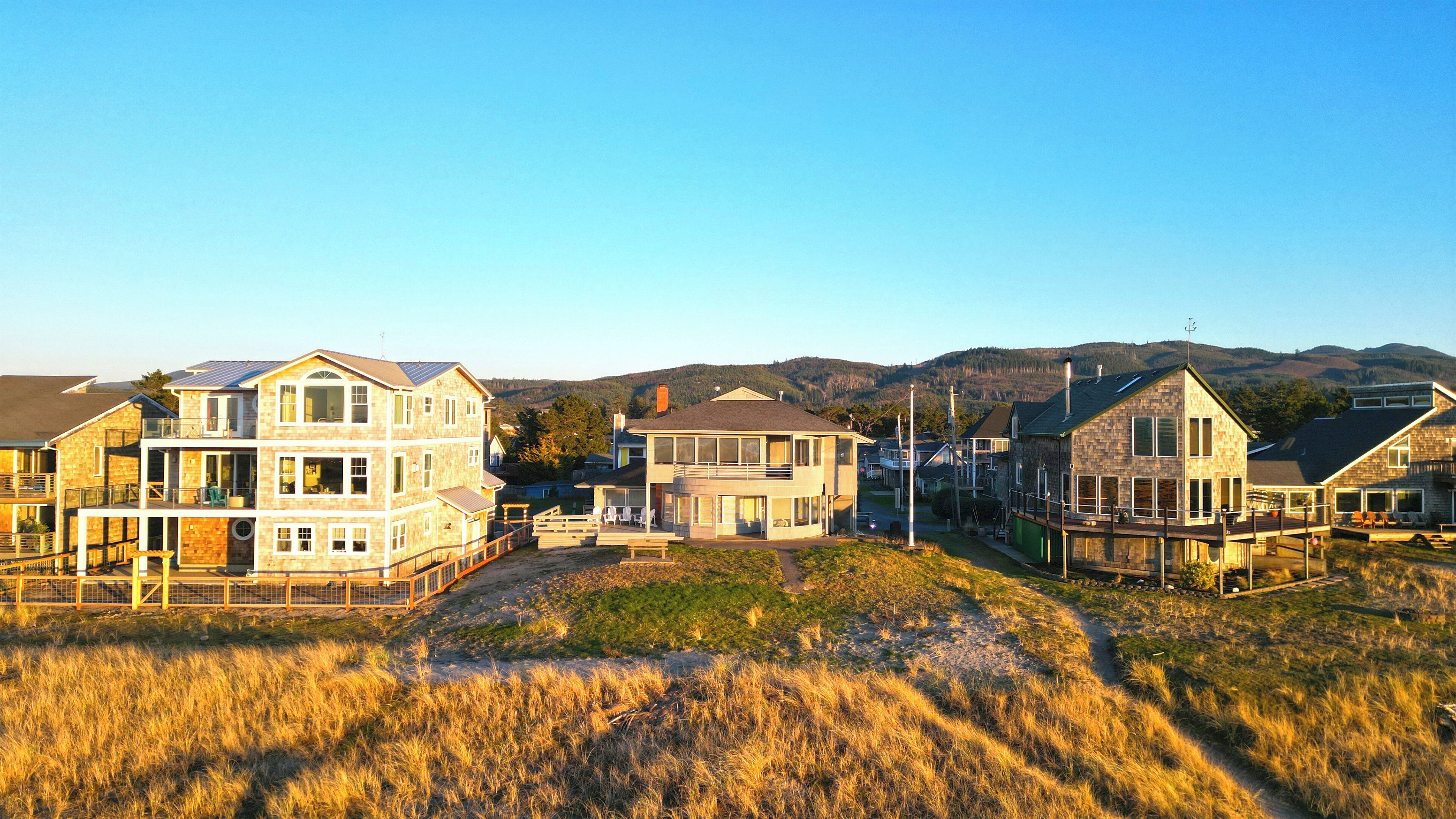 A coastal residential area with multi-story houses, a grassy foreground, and a mountainous backdrop.