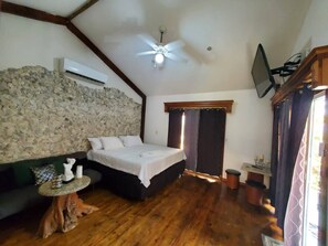 Un dormitorio con pared de piedra, suelo de madera, cama, mesita de noche y ventilador de techo.