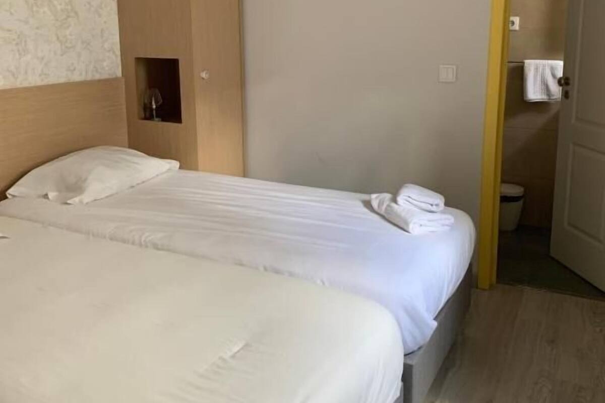 Une chambre d’hôtel avec deux lits, une armoire en bois, une salle de bain visible à travers une porte ouverte et une petite étagère avec une lampe.