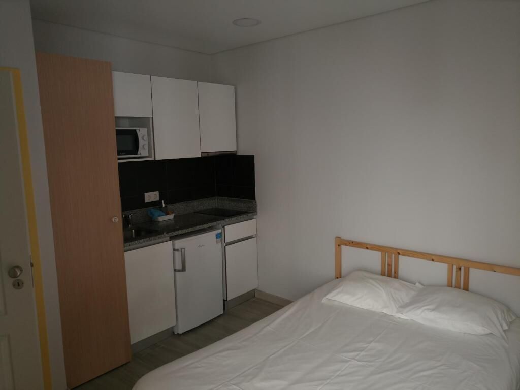 A small room with a bed, a kitchenette with a microwave, and a wooden door.