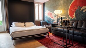 A hotel room with a bed, a sofa, a table with bottles, and a large wall painting. - DECA CONCIERGERIE (Le Muy)
