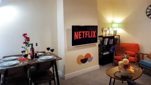 A living room with a dining area, a bookshelf, and a wall-mounted TV displaying 'NETFLIX'. - Near Uni and Hospital - Long Stays - Fast Wi-Fi - 15 Mins to City (Manchester)