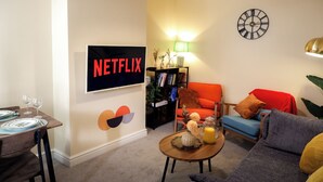 A living room with a TV displaying 'NETFLIX', a bookshelf, a coffee table with decorative items, and a couch. - Near Uni and Hospital - Long Stays - Fast Wi-Fi - 15 Mins to City (Manchester)