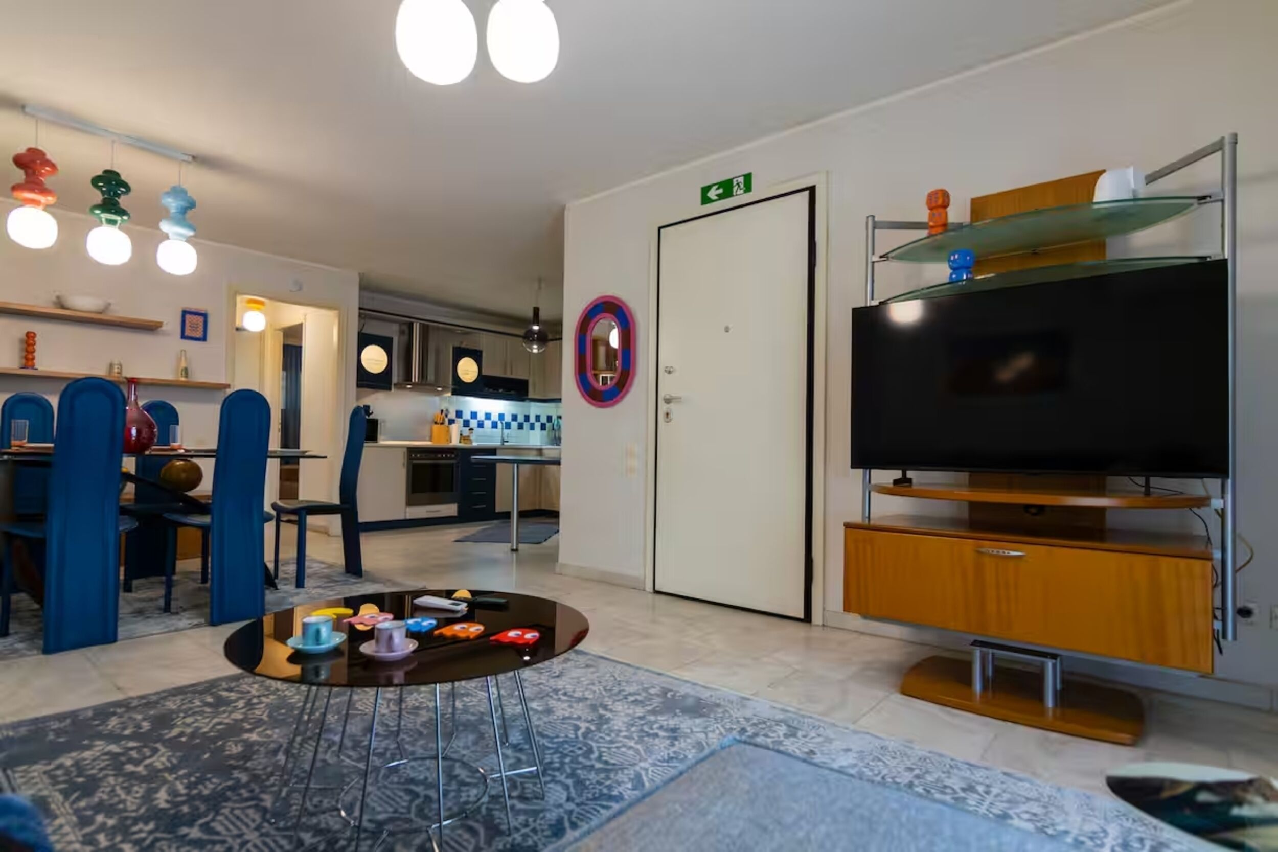 Een moderne woonkamer met een televisie, een salontafel met speelgoed, een eethoek met blauwe stoelen en een keuken op de achtergrond.