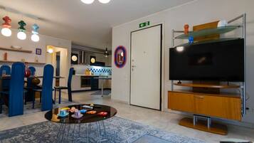 Een moderne woonkamer met een televisie, een salontafel met speelgoed, een eethoek met blauwe stoelen en een keuken op de achtergrond.