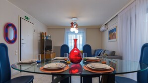 Een eethoek met een glazen tafel, blauwe stoelen, een rode vaas en een televisie op de achtergrond.