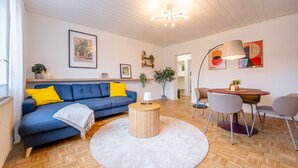 A living room with a blue sofa, a round wooden coffee table, a white rug, a dining area with a table and chairs, and a ceiling light fixture. - New Apartment, 5 Minutes to Centro and Gasometer (Oberhausen)