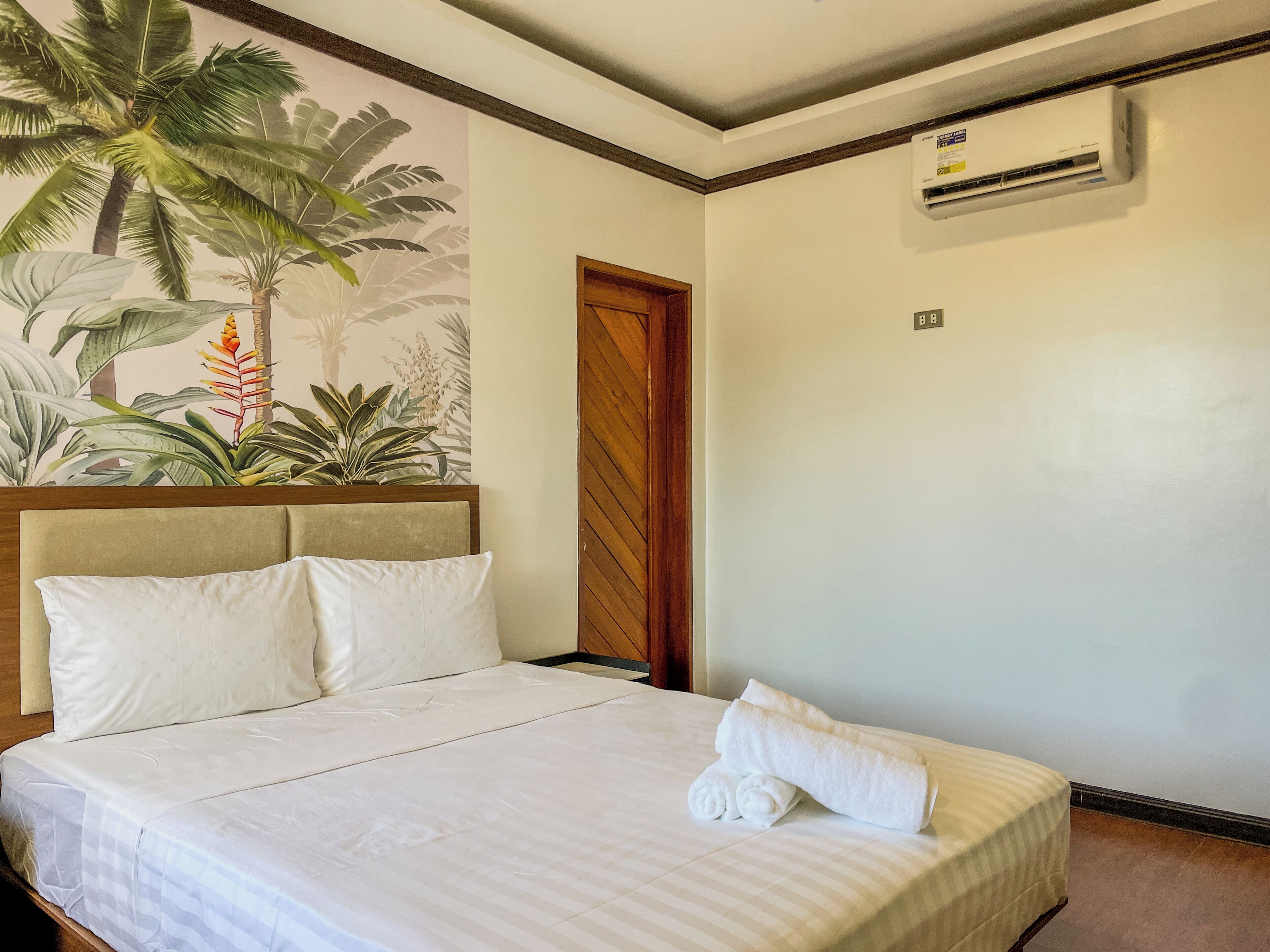 Een net opgemaakt bed met witte lakens en kussens, een tropische muurschildering, een houten deur en een wandairconditioner.