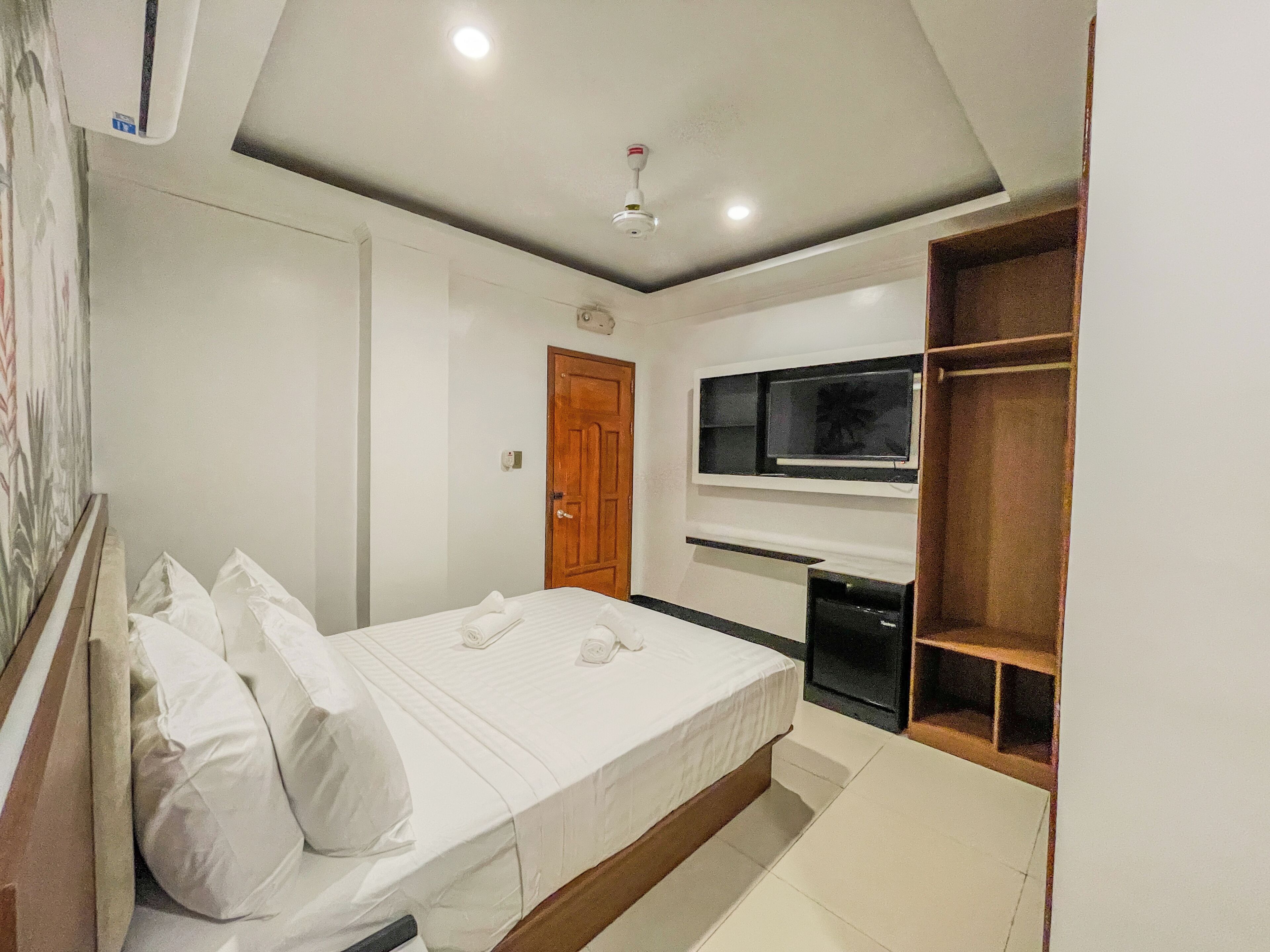 A bedroom with a bed, a TV mounted on the wall, a door, and a small shelf.