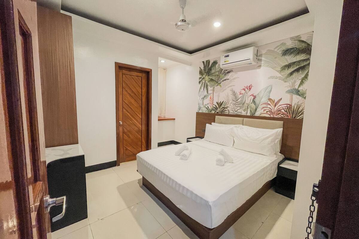 Habitación de hotel con cama, puerta de madera, unidad de aire acondicionado y un mural temático tropical.