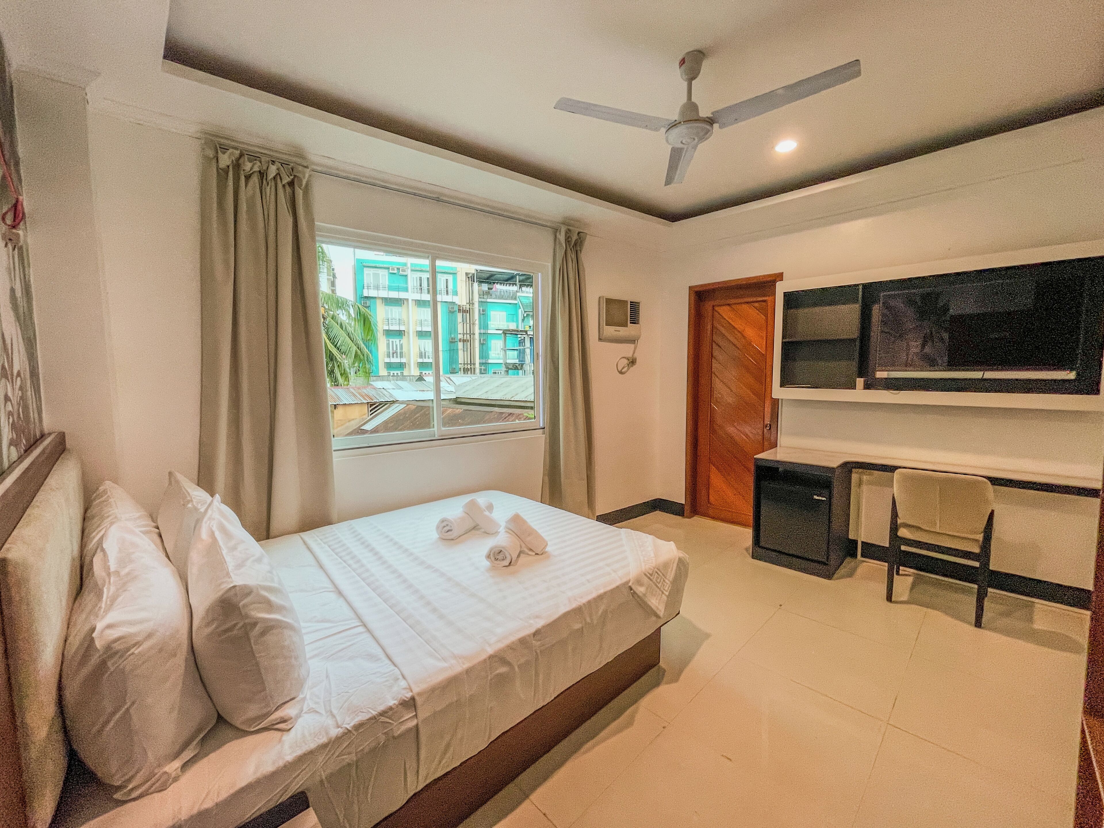 Une chambre d’hôtel équipée d’un lit, d’un bureau, d’une chaise, d’un ventilateur de plafond, d’une fenêtre avec des rideaux et d’un téléviseur fixé au mur.