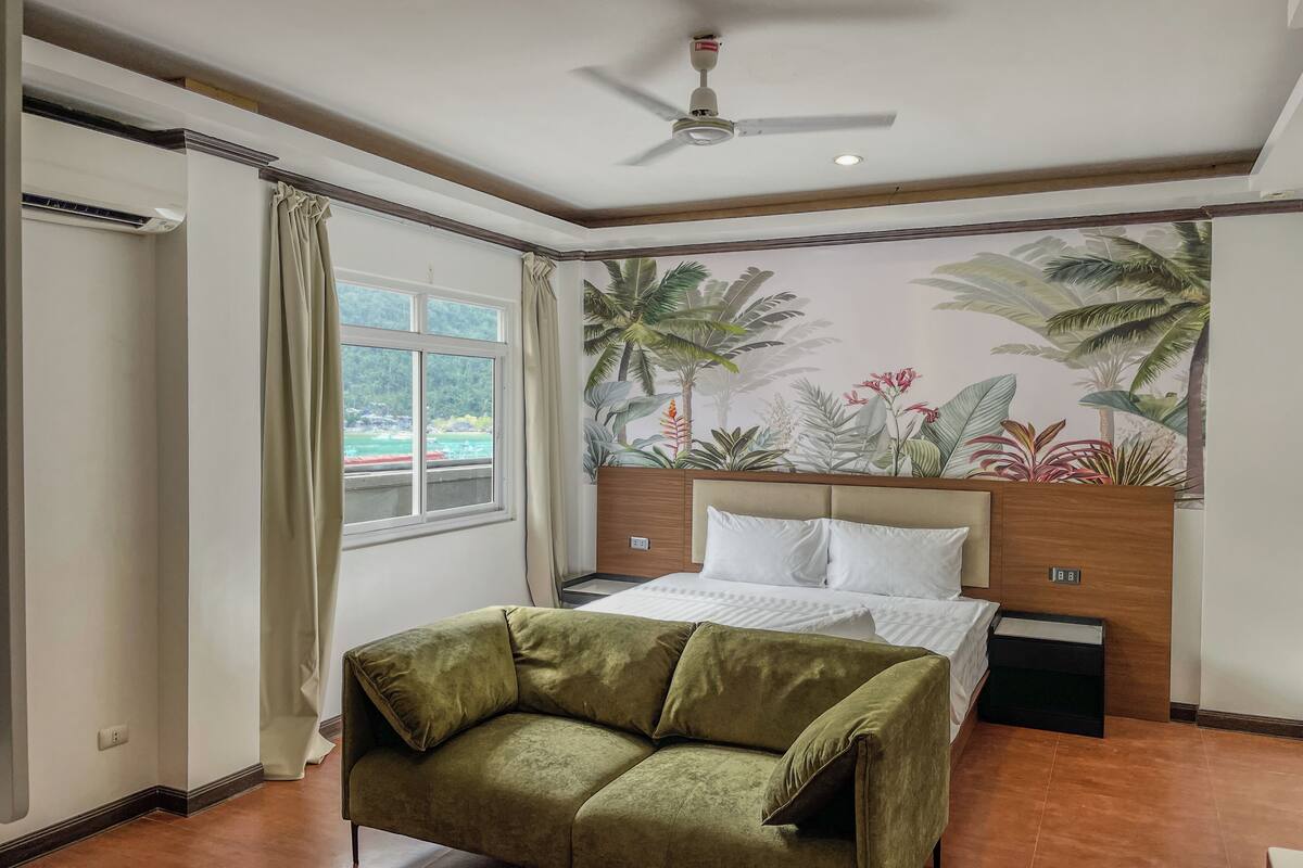 Un dormitorio con una cama, un sofá verde, un ventilador de techo, una ventana con cortinas y un mural tropical en la pared.