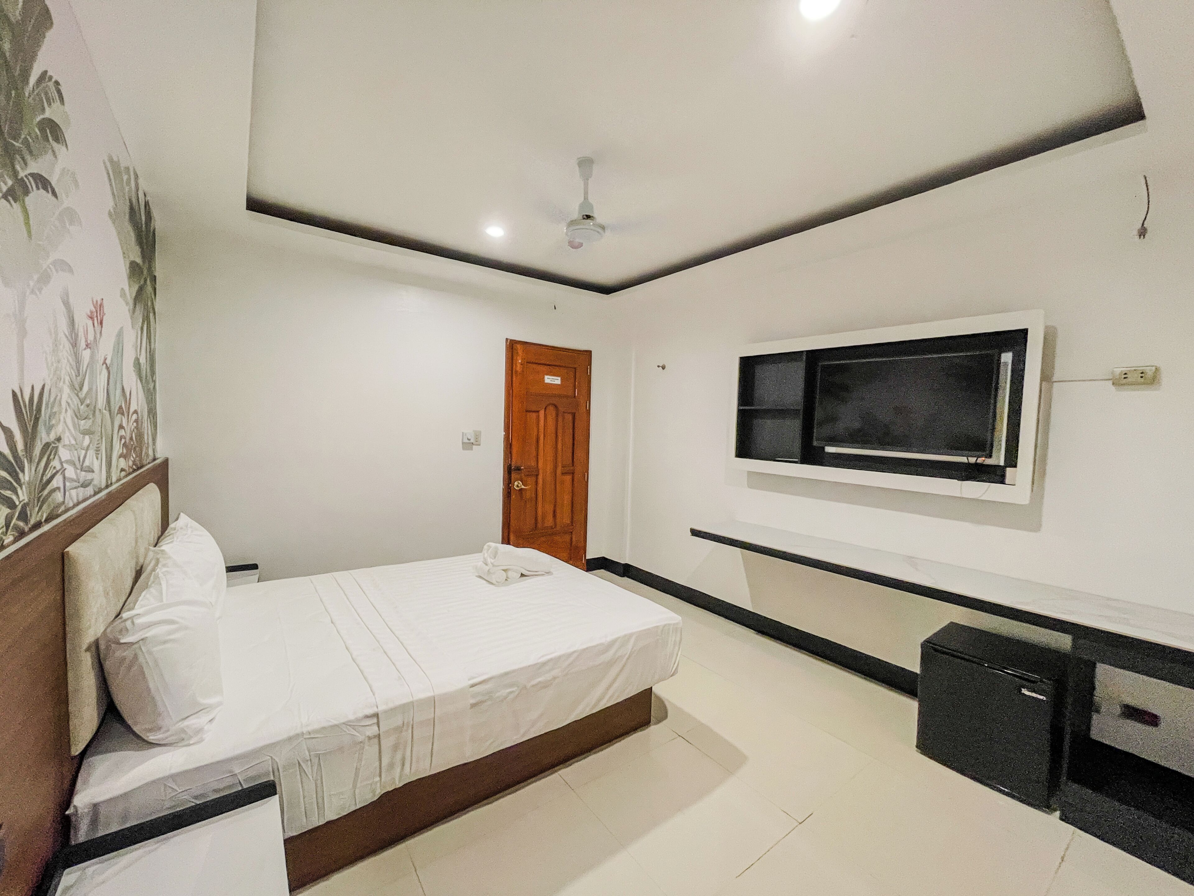 Une chambre à coucher comprenant un lit, une porte, un ventilateur de plafond, un téléviseur fixé au mur et un petit meuble noir.