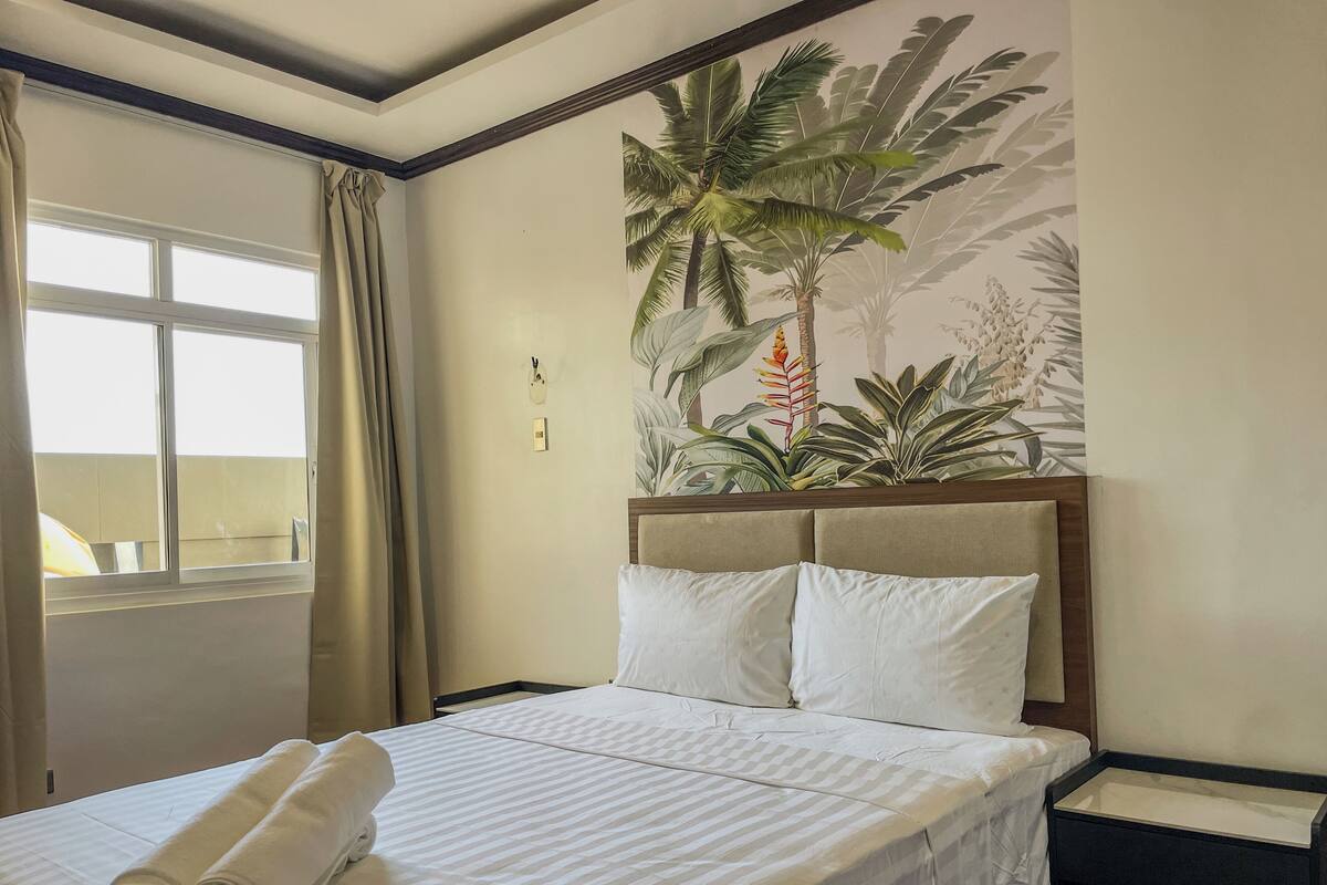 Una habitación de hotel con una cama, una ventana con cortinas, un mural tropical en la pared y una mesita de noche.