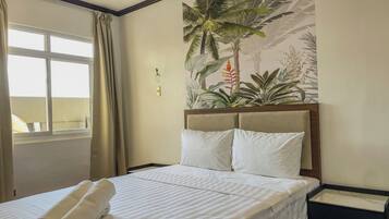 Una habitación de hotel con una cama, una ventana con cortinas, un mural tropical en la pared y una mesita de noche.