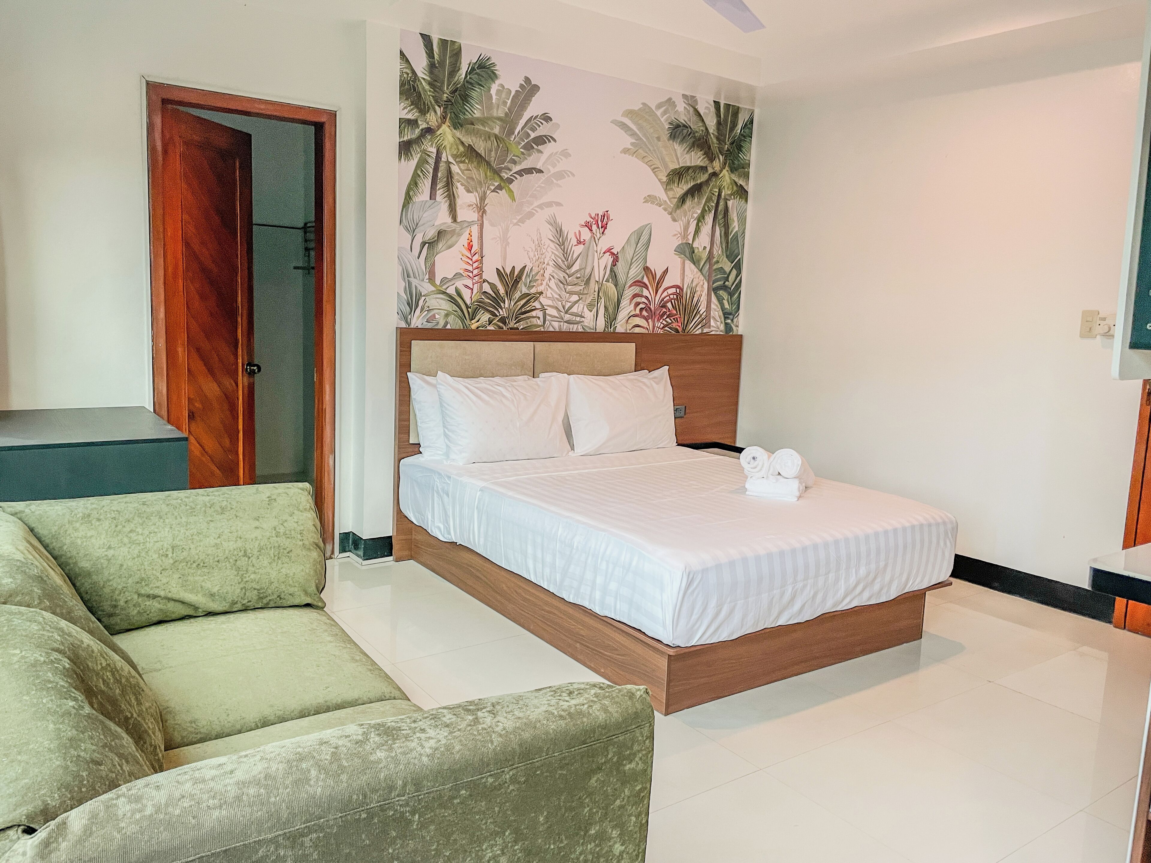 A bedroom with a bed, a green sofa, a wooden door, and a wall with a tropical plant mural.