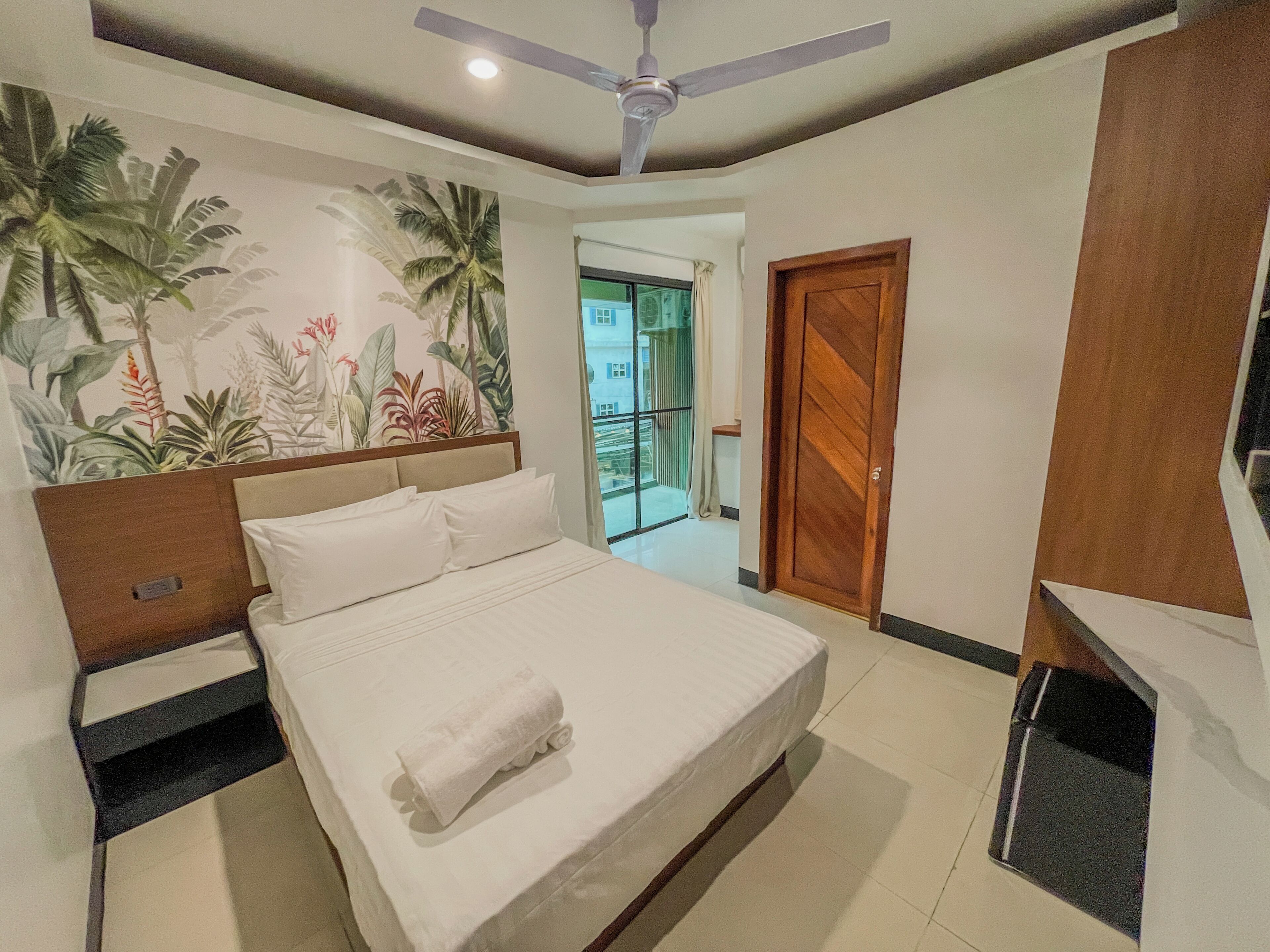 A hotel room with a bed, a ceiling fan, a nightstand, a wooden door, and a view of the outside.