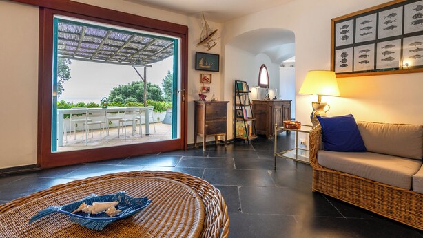 A spacious living area with a wicker sofa, a wicker chair, a dining table with chairs, a glass coffee table, and a large window with a view of the outdoors. - Relax e Natura a Punta Rossa (San Felice Circeo)