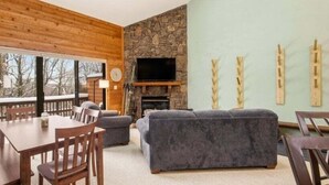 Un salon chaleureux avec une cheminée en pierre, des murs en bois, une grande fenêtre et un coin repas comprenant une table et des chaises.