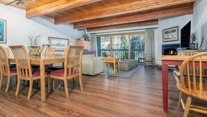 A spacious living room with wooden floors, a dining area with wicker chairs, a fireplace, and a large window with a view of trees. - Riverside Retreat 2 Bedroom Condo (Telluride)