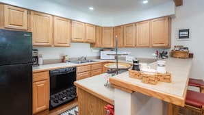A kitchen with wooden cabinets, a black refrigerator, a dishwasher, a sink, and a small island with stools. - Riverside Retreat 2 Bedroom Condo (Telluride)