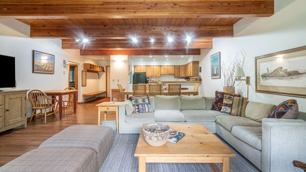 A cozy living room with a sofa, a coffee table, and a TV. There is a kitchen in the background with wooden cabinets and a dining area with chairs. - Riverside Retreat 2 Bedroom Condo (Telluride)