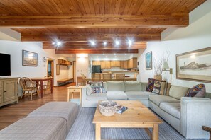 A cozy living room with a sofa, a coffee table, and a TV. There is a kitchen in the background with wooden cabinets and a dining area with chairs. - Riverside Retreat 2 Bedroom Condo (Telluride)